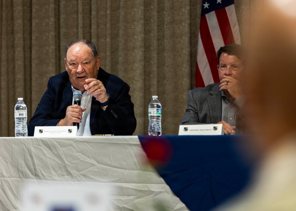 Two men are seated at a panel table during what appears to be a formal event or conference. The older man on the left is speaking into a microphone while the younger man on the right listens attentively. An American flag is visible in the background, and name placards are placed on the table in front of them.