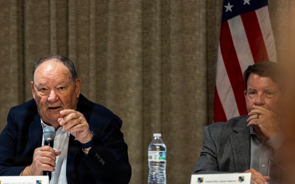 Two men are seated at a panel table during what appears to be a formal event or conference. The older man on the left is speaking into a microphone while the younger man on the right listens attentively. An American flag is visible in the background, and name placards are placed on the table in front of them.
