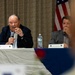 Two men are seated at a panel table during what appears to be a formal event or conference. The older man on the left is speaking into a microphone while the younger man on the right listens attentively. An American flag is visible in the background, and name placards are placed on the table in front of them.