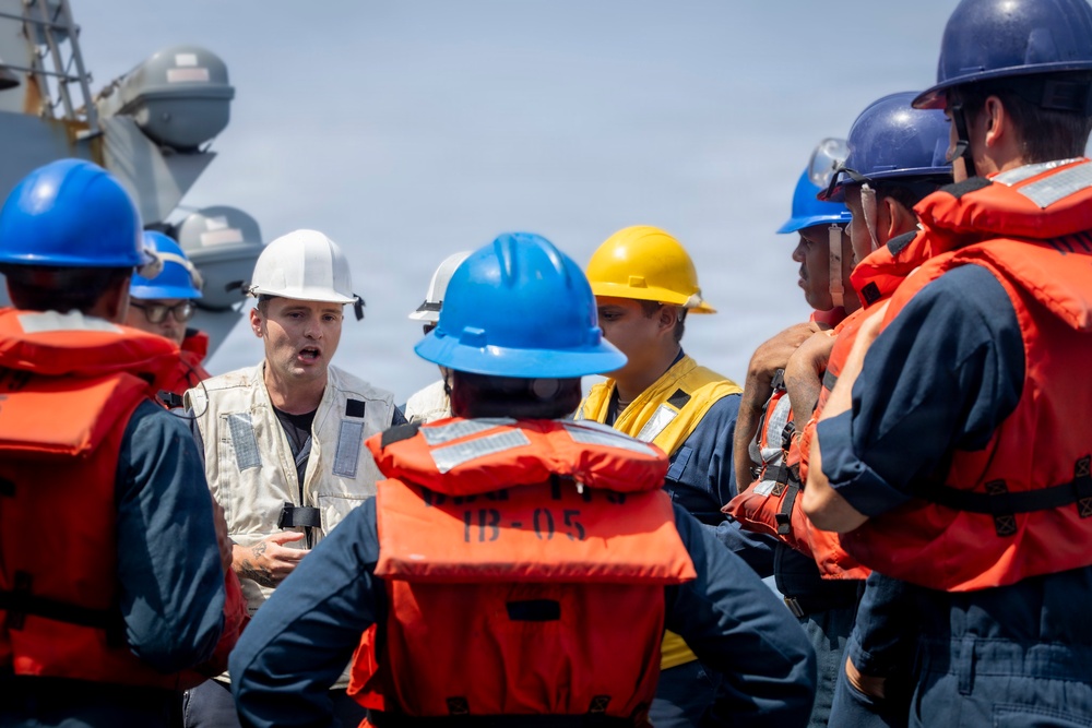 A group of personnel wearing hard hats and orange life vests are gathered on the deck of a naval vessel for what appears to be a briefing or safety drill. A man in a white hard hat and beige vest, holding binoculars, appears to be the instructor or supervisor addressing the group. Naval ship structures are visible in the background.