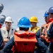 A group of personnel wearing hard hats and orange life vests are gathered on the deck of a naval vessel for what appears to be a briefing or safety drill. A man in a white hard hat and beige vest, holding binoculars, appears to be the instructor or supervisor addressing the group. Naval ship structures are visible in the background.