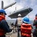 Naval personnel are conducting a small rigid inflatable boat (RIB) hoisting operation on the deck of a warship at sea. Four crew members wearing orange life vests and blue hard hats are managing lines and overseeing the crane lift, while one supervisor in a white hard hat and gray vest labeled 'BMC' observes. The operation appears to be a routine small boat recovery or deployment exercise.