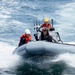 Three personnel are operating a gray rigid inflatable boat (RIB) at speed on open ocean waters. The crew members are wearing safety equipment including helmets and life vests, suggesting a military or coast guard operation. The boat is equipped with communications equipment including radio antennas and a console-mounted display.