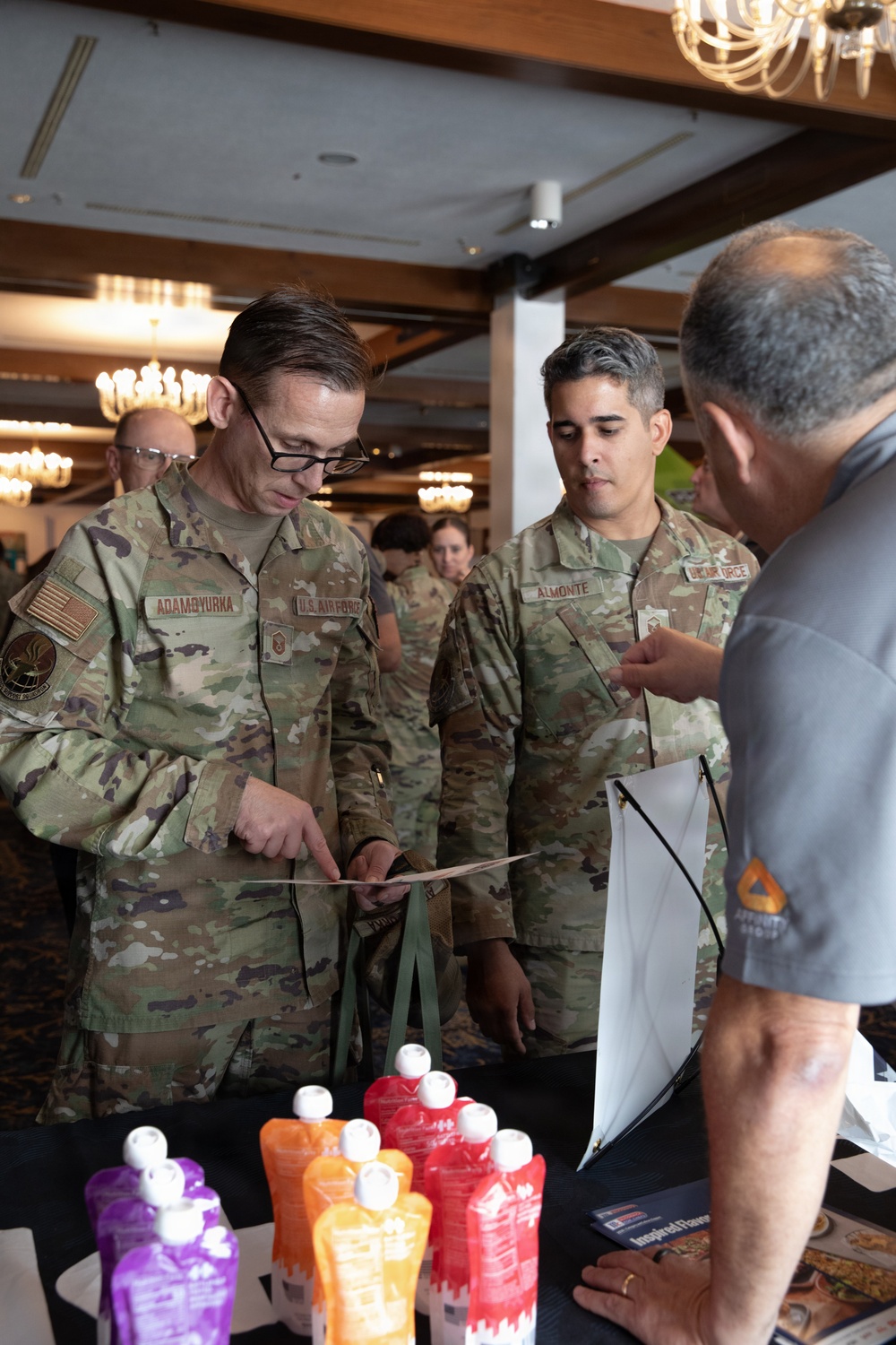 Two U.S. Air Force personnel in OCP (Operational Camouflage Pattern) uniforms are examining materials at a vendor or exhibition booth. The service members, identified by name tapes as 'ADAMOYURKA' and 'ALMONTE,' are interacting with a civilian representative. The setting appears to be an indoor conference or trade show event in a hotel ballroom.