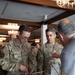 Two U.S. Air Force personnel in OCP (Operational Camouflage Pattern) uniforms are examining materials at a vendor or exhibition booth. The service members, identified by name tapes as 'ADAMOYURKA' and 'ALMONTE,' are interacting with a civilian representative. The setting appears to be an indoor conference or trade show event in a hotel ballroom.