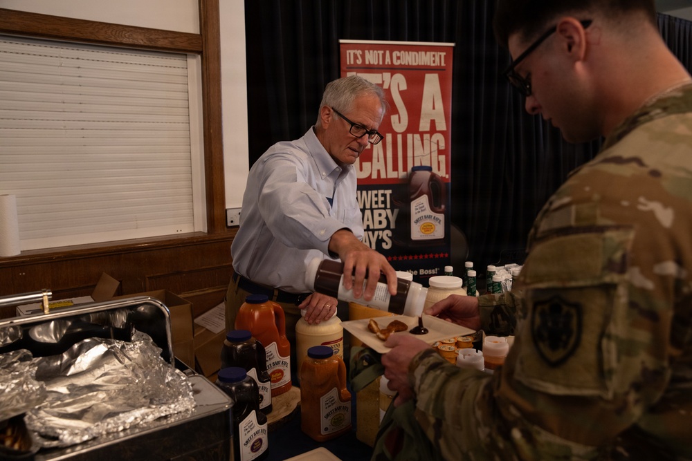 A civilian man in a white dress shirt is serving or demonstrating Sweet Baby Ray's barbecue sauce products to a uniformed military service member at what appears to be a food vendor booth or tasting event. The setting looks like an indoor venue with a promotional banner for Sweet Baby Ray's visible in the background. Multiple sauce bottles and food items are spread across the table.