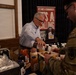 A civilian man in a white dress shirt is serving or demonstrating Sweet Baby Ray's barbecue sauce products to a uniformed military service member at what appears to be a food vendor booth or tasting event. The setting looks like an indoor venue with a promotional banner for Sweet Baby Ray's visible in the background. Multiple sauce bottles and food items are spread across the table.