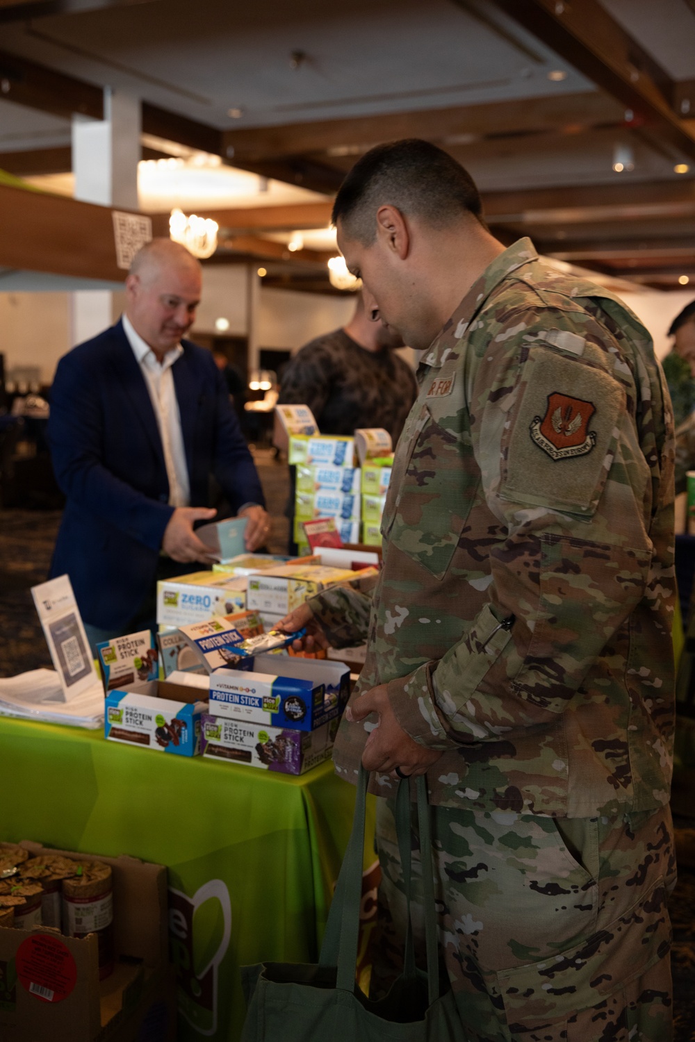 A US Air Force service member in OCP (Operational Camouflage Pattern) uniform is examining products at a vendor booth during what appears to be a health and wellness expo or military trade show. The booth displays protein sticks, zero-sugar snacks, and other nutritional products on a green-tablecloth-covered table. A civilian man in a blue blazer stands across the table, likely a vendor representative.