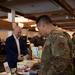 A US Air Force service member in OCP (Operational Camouflage Pattern) uniform is examining products at a vendor booth during what appears to be a health and wellness expo or military trade show. The booth displays protein sticks, zero-sugar snacks, and other nutritional products on a green-tablecloth-covered table. A civilian man in a blue blazer stands across the table, likely a vendor representative.