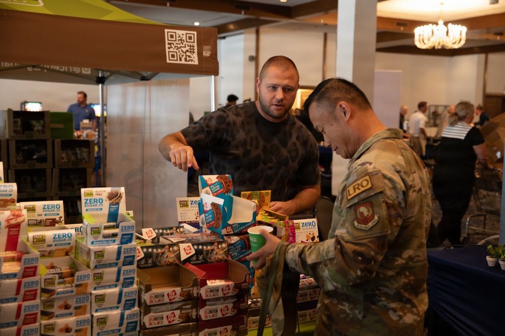 A vendor in a camouflage t-shirt is presenting food products to a military personnel wearing a multicam uniform with an 'LRO' patch at what appears to be a trade show or military expo. The table is stacked with numerous boxes of 'Zero Sugar' snack bars and protein products. The event is taking place in a large indoor venue with other booths and attendees visible in the background.