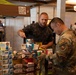 A vendor in a camouflage t-shirt is presenting food products to a military personnel wearing a multicam uniform with an 'LRO' patch at what appears to be a trade show or military expo. The table is stacked with numerous boxes of 'Zero Sugar' snack bars and protein products. The event is taking place in a large indoor venue with other booths and attendees visible in the background.