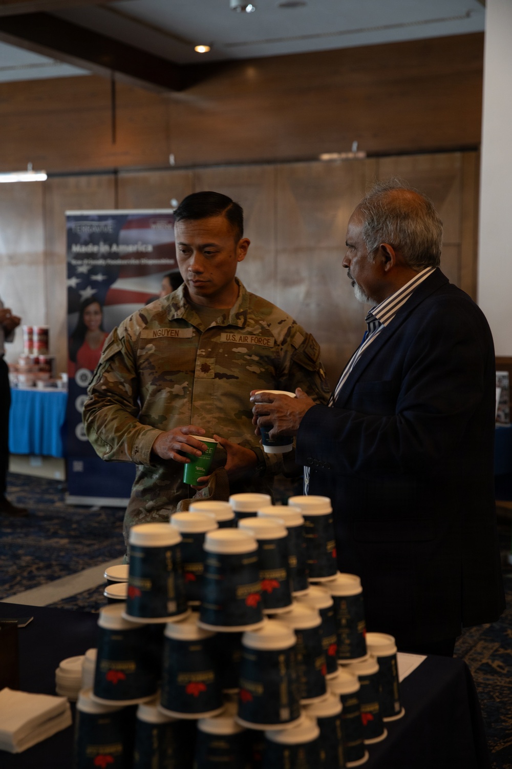 A U.S. Air Force servicemember named Nguyen is engaged in conversation with a civilian man at what appears to be a vendor expo or trade show event. Both individuals are holding small product containers, with a display table in the foreground stacked with dark blue lidded cups or containers. A banner in the background reads 'Made in America' with an American flag design.