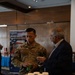 A U.S. Air Force servicemember named Nguyen is engaged in conversation with a civilian man at what appears to be a vendor expo or trade show event. Both individuals are holding small product containers, with a display table in the foreground stacked with dark blue lidded cups or containers. A banner in the background reads 'Made in America' with an American flag design.