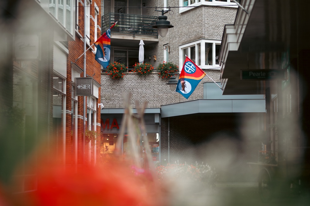 A street scene in what appears to be a European city, showing a mix of brick buildings with balconies decorated with red flowering plants. Two colorful flags are prominently displayed on the building facade, featuring blue, red, and multicolored designs with emblems. A street sign reading 'Pearle' is visible on the right side, and various shop signs including '+SIER' and partial letters 'MA' are visible.