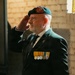An older male veteran is photographed in profile while performing a formal salute. He is wearing a teal/green beret with a red badge, a dark blazer, and a row of service medals on his chest. The setting appears to be an indoor ceremony or commemorative event.