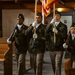 A military color guard of four soldiers in U.S. Army dress green uniforms is marching in formation inside what appears to be a chapel or church setting. Two soldiers carry flags (including a U.S. flag) while two others carry rifles at ceremonial positions. The setting suggests a formal military ceremony or memorial service.