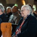 An elderly woman with white hair and glasses sits in a church pew, wearing a dark blazer and a red, white, and blue scarf or lanyard. She appears to be attending a formal or commemorative gathering, possibly a memorial or ceremony, based on the setting and attire. A name badge reading 'Jeanne' is visible on her lanyard.
