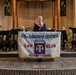 A woman speaks at a podium during a formal ceremony inside what appears to be a church or chapel. A banner in the foreground reads '82nd Airborne Division C47 Club Holland Chapter,' indicating a commemorative or memorial event. A band or ensemble dressed in black is seated behind her, suggesting a musical component to the ceremony.