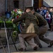 Two individuals in olive/khaki military-style uniforms are placing a floral wreath on a memorial stand during what appears to be a formal commemoration ceremony. The wreath features red, white, and blue flowers with matching ribbons, suggesting a national or military memorial event. A crowd of onlookers, some photographing the ceremony, observes from the background near a brick building.