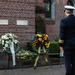 A uniformed military or naval officer stands with his back to the camera, facing three memorial wreaths displayed on stands against a brick building. The wreaths include one with red, white, and blue ribbons suggesting American affiliation, one with white flowers and a white ribbon, and one with yellow sunflowers and a gold ribbon. The scene depicts a formal wreath-laying or commemorative ceremony.