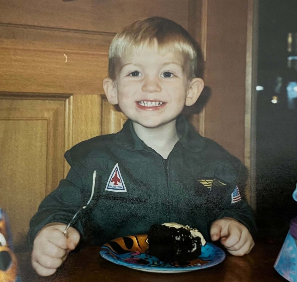 A young blonde boy, approximately 3-5 years old, is smiling broadly at the camera while sitting at a table with a slice of chocolate cake on a decorative plate in front of him. He is wearing a dark olive/green zip-up jacket with military-style patches, including one labeled 'PHANTOM' with a jet aircraft graphic and a small American flag patch. The photo appears to be a scanned print photograph, likely from the late 1990s to early 2000s based on image quality and style.