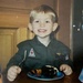 A young blonde boy, approximately 3-5 years old, is smiling broadly at the camera while sitting at a table with a slice of chocolate cake on a decorative plate in front of him. He is wearing a dark olive/green zip-up jacket with military-style patches, including one labeled 'PHANTOM' with a jet aircraft graphic and a small American flag patch. The photo appears to be a scanned print photograph, likely from the late 1990s to early 2000s based on image quality and style.