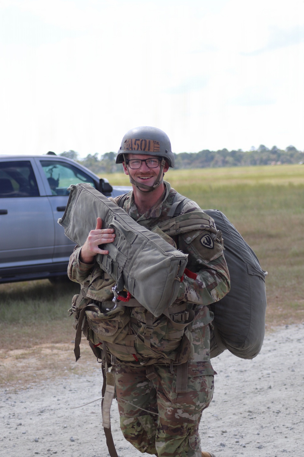 A smiling male soldier in U.S. Army OCP (Operational Camouflage Pattern) uniform is carrying parachute equipment after what appears to be a airborne jump operation. He is wearing a PASGT or ACH helmet with a name/unit tape visible, glasses, and is loaded with a parachute pack tray and reserve parachute. The setting appears to be a drop zone or landing zone at a military installation.