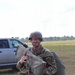 A smiling male soldier in U.S. Army OCP (Operational Camouflage Pattern) uniform is carrying parachute equipment after what appears to be a airborne jump operation. He is wearing a PASGT or ACH helmet with a name/unit tape visible, glasses, and is loaded with a parachute pack tray and reserve parachute. The setting appears to be a drop zone or landing zone at a military installation.