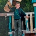 A young boy stands on an elevated wooden playground structure, smiling at the camera. He is holding a homemade cardboard parachute or glider above his head with one hand while wearing a backpack. The photo is dated August 20, 2007, based on the timestamp visible in the lower right corner.