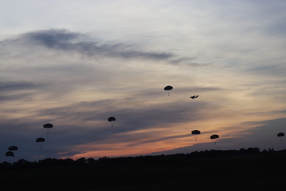 A twilight/dusk military airborne operation showing multiple parachutists descending against a dramatic sunset sky with orange and purple hues. A military transport aircraft is visible in the upper right portion of the frame, having recently deployed the paratroopers. The silhouetted treeline at the bottom provides a dark contrast to the colorful sky.