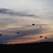 A twilight/dusk military airborne operation showing multiple parachutists descending against a dramatic sunset sky with orange and purple hues. A military transport aircraft is visible in the upper right portion of the frame, having recently deployed the paratroopers. The silhouetted treeline at the bottom provides a dark contrast to the colorful sky.