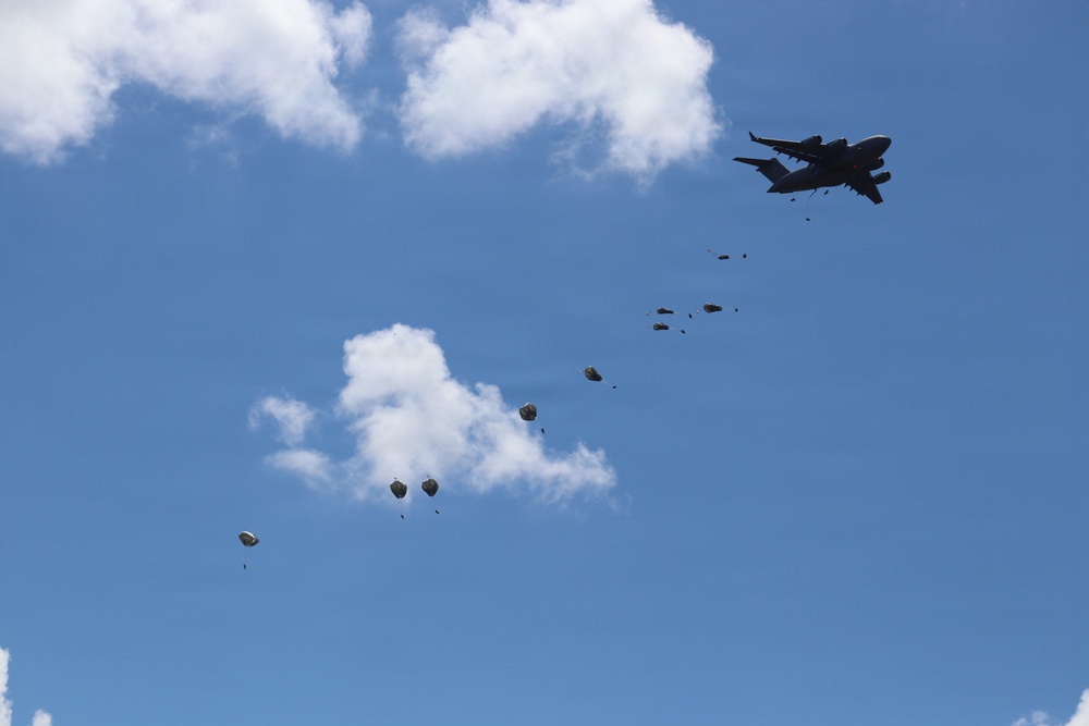 A large military transport aircraft is conducting an aerial airdrop operation against a blue sky with scattered white clouds. Multiple cargo bundles at various stages of descent are visible, with some parachutes already deployed on the lower-left bundles and others still in free fall closer to the aircraft. The trail of bundles creates a diagonal line across the frame from upper-right to lower-left.