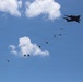 A large military transport aircraft is conducting an aerial airdrop operation against a blue sky with scattered white clouds. Multiple cargo bundles at various stages of descent are visible, with some parachutes already deployed on the lower-left bundles and others still in free fall closer to the aircraft. The trail of bundles creates a diagonal line across the frame from upper-right to lower-left.