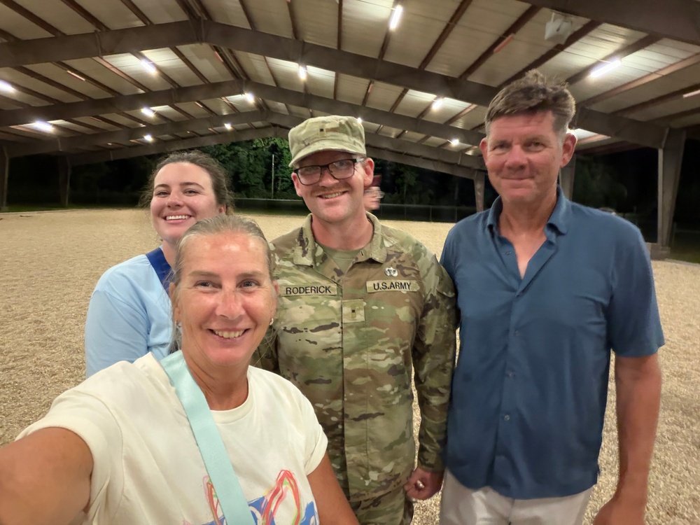 A group of four people posing for a selfie inside what appears to be an open-air covered arena or pavilion at night. One individual is wearing a U.S. Army uniform with the name tape 'RODERICK' and an OCP (Operational Camouflage Pattern) uniform. The group appears to be in a casual, celebratory gathering.