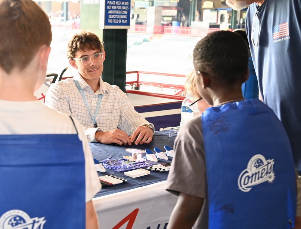A young man wearing glasses and a lanyard sits behind a table at what appears to be a STEM or educational demonstration booth inside a baseball stadium. He is engaging with two children wearing blue 'Comets' vests, while an older adult stands nearby. Various small electronic or science components are displayed on the table.