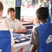 A young man wearing glasses and a lanyard sits behind a table at what appears to be a STEM or educational demonstration booth inside a baseball stadium. He is engaging with two children wearing blue 'Comets' vests, while an older adult stands nearby. Various small electronic or science components are displayed on the table.