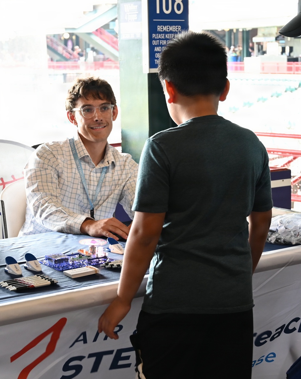 A young adult male volunteer or representative sits behind a display table at what appears to be a STEM outreach event inside a baseball stadium. He is engaging with a young boy who is standing across the table, examining electronic circuit board displays and educational materials. The event appears to be sponsored by an organization with 'AIR' and 'STEM' visible on the table banner.