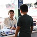 A young adult male volunteer or representative sits behind a display table at what appears to be a STEM outreach event inside a baseball stadium. He is engaging with a young boy who is standing across the table, examining electronic circuit board displays and educational materials. The event appears to be sponsored by an organization with 'AIR' and 'STEM' visible on the table banner.