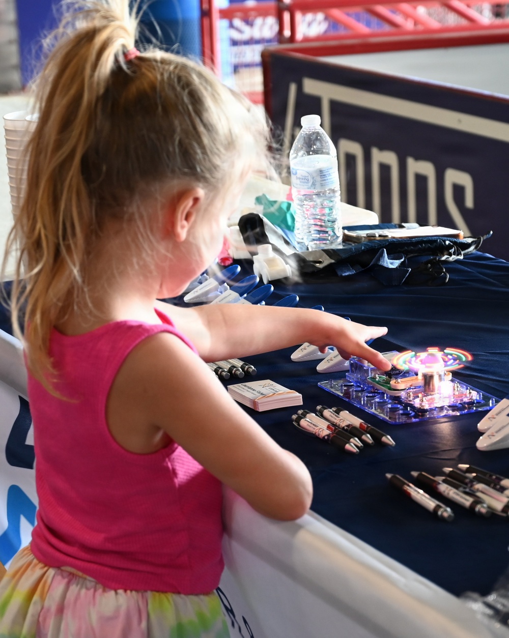 A young blonde girl in a pink tank top reaches toward a glowing LED spinner toy on a navy blue promotional table. The table appears to be a vendor or sponsor booth, likely at a sporting event, displaying pens, business cards, and branded materials. The backdrop includes partial text reading 'Tonos' or similar branding on the navy tablecloth.
