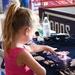 A young blonde girl in a pink tank top reaches toward a glowing LED spinner toy on a navy blue promotional table. The table appears to be a vendor or sponsor booth, likely at a sporting event, displaying pens, business cards, and branded materials. The backdrop includes partial text reading 'Tonos' or similar branding on the navy tablecloth.