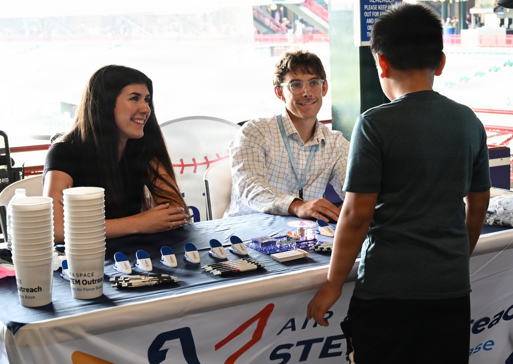 Two representatives, a woman and a man wearing a lanyard, are staffing an Air Force STEM Outreach booth at what appears to be a baseball stadium venue. They are smiling and engaging with a young boy who has approached their table. The table displays promotional items including pens, small blue items, cups, and electronic components.