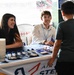 Two representatives, a woman and a man wearing a lanyard, are staffing an Air Force STEM Outreach booth at what appears to be a baseball stadium venue. They are smiling and engaging with a young boy who has approached their table. The table displays promotional items including pens, small blue items, cups, and electronic components.