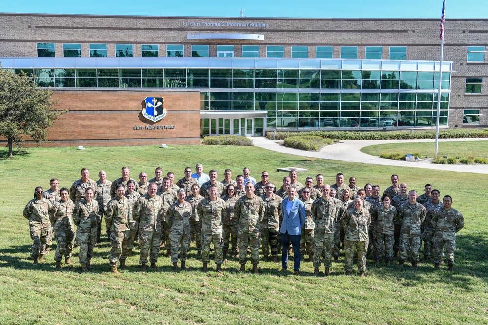 A large group of military personnel, predominantly in camouflage uniforms, pose for a formal group photograph on a grass lawn in front of the 557th Weather Wing headquarters building. The building is identified as the 'Lt. Gen. Thomas S. Moorman Building' and displays the 557th Weather Wing emblem on its brick facade. One individual in a light blue civilian suit stands prominently in the center of the group.