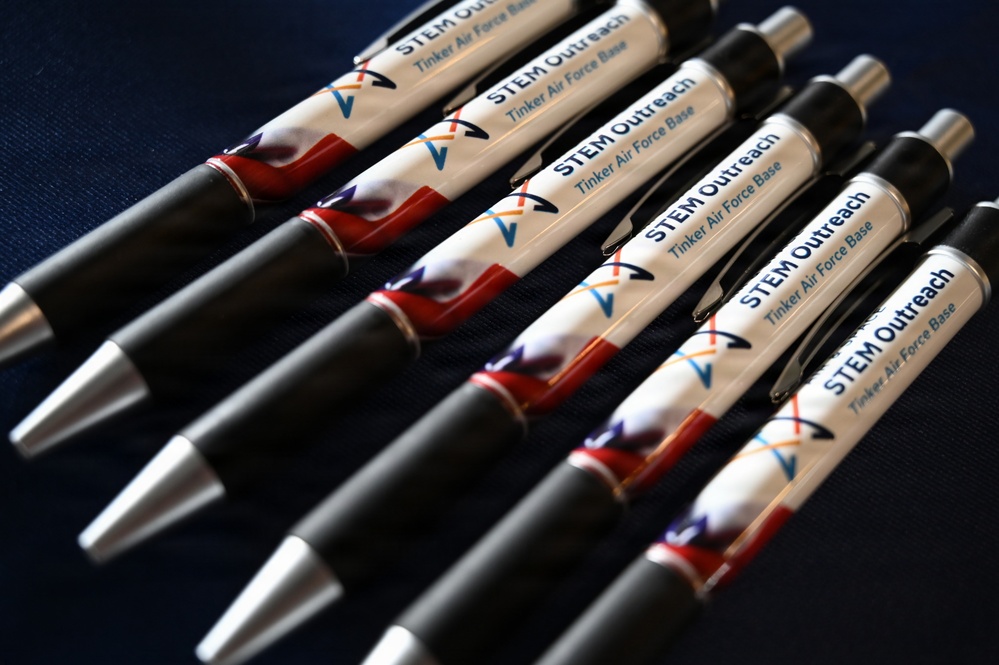 The image shows a collection of promotional ballpoint pens arranged in a fan-like pattern on a dark blue surface. Each pen is branded with 'STEM Outreach' and 'Tinker Air Force Base' text, featuring a patriotic red, white, and blue design with a logo consisting of intersecting arrows or chevrons. These appear to be branded merchandise items likely distributed at outreach or recruitment events.