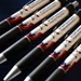 The image shows a collection of promotional ballpoint pens arranged in a fan-like pattern on a dark blue surface. Each pen is branded with 'STEM Outreach' and 'Tinker Air Force Base' text, featuring a patriotic red, white, and blue design with a logo consisting of intersecting arrows or chevrons. These appear to be branded merchandise items likely distributed at outreach or recruitment events.