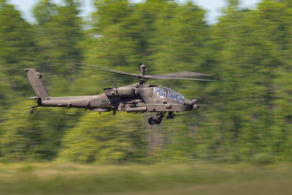 A U.S. Army attack helicopter, identifiable as an AH-64 Apache, is captured in low-level flight at high speed against a blurred green tree line background. The motion blur on the background indicates the use of a panning photography technique, keeping the aircraft sharp while conveying a strong sense of speed. The helicopter appears to be in an operational or training flight configuration.