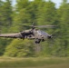 A U.S. Army attack helicopter, identifiable as an AH-64 Apache, is captured in low-level flight at high speed against a blurred green tree line background. The motion blur on the background indicates the use of a panning photography technique, keeping the aircraft sharp while conveying a strong sense of speed. The helicopter appears to be in an operational or training flight configuration.