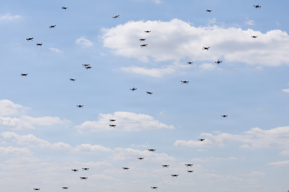 The image shows a large swarm of multirotor drones (quadcopters or hexacopters) flying in a loosely distributed formation against a partly cloudy blue sky. The drones appear to be small consumer or commercial-grade UAVs, visible as dark silhouettes at varying altitudes and distances. This appears to be a coordinated drone swarm demonstration or show.