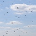 The image shows a large swarm of multirotor drones (quadcopters or hexacopters) flying in a loosely distributed formation against a partly cloudy blue sky. The drones appear to be small consumer or commercial-grade UAVs, visible as dark silhouettes at varying altitudes and distances. This appears to be a coordinated drone swarm demonstration or show.