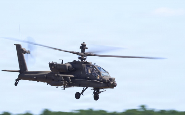 A military attack helicopter, consistent with an AH-64 Apache design, is captured in low-level flight against a light blue sky with a blurred tree line in the background. The motion blur on the rotor blades and background indicates the helicopter is moving at speed. The image appears to be taken with a panning technique, keeping the aircraft sharp while blurring the surroundings.
